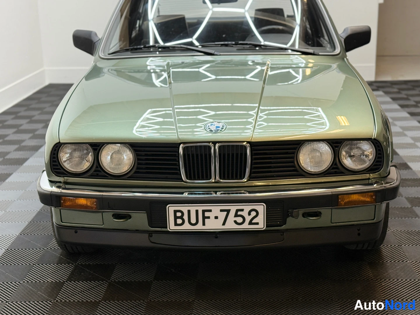 Edestä: BMW 324d 1986, parked, head-on exterior shot, licence plate, round headlights and chrome kidney grille, light green metallic paint, reflectance of indoor ceilin