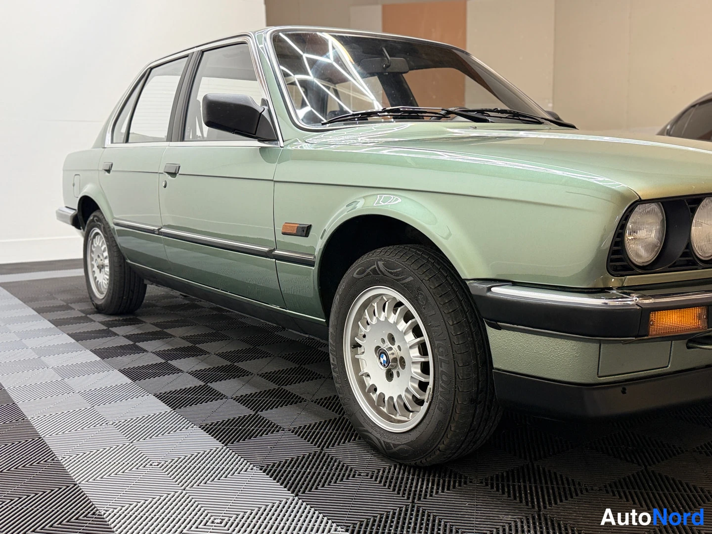 Etuviisto, nokka oikealle: BMW 324d 1986, parked indoor space, angled exterior shot showing grille and left flank, alloy wheels, side indicator, chrome trim, light green metallic p