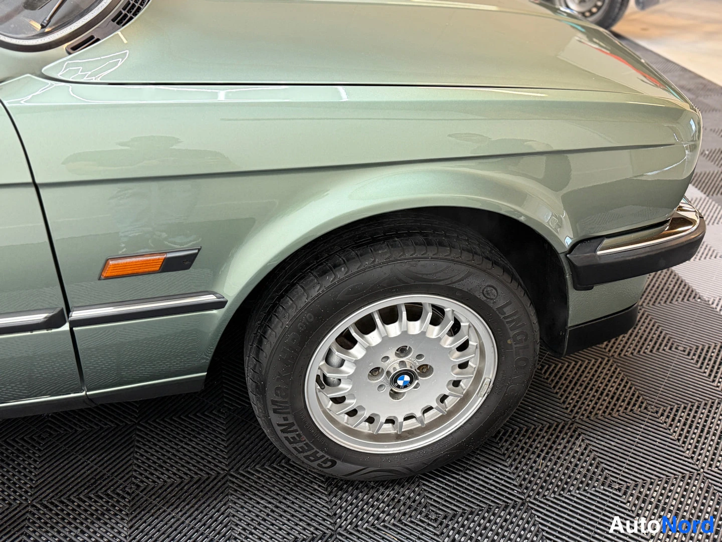Etuviisto, nokka oikealle: BMW 324d 1986, close-up of left flank and front fender, alloy wheel with BMW hubcap and Dunlop tyre, side indicator, light green metallic paint, indoor c