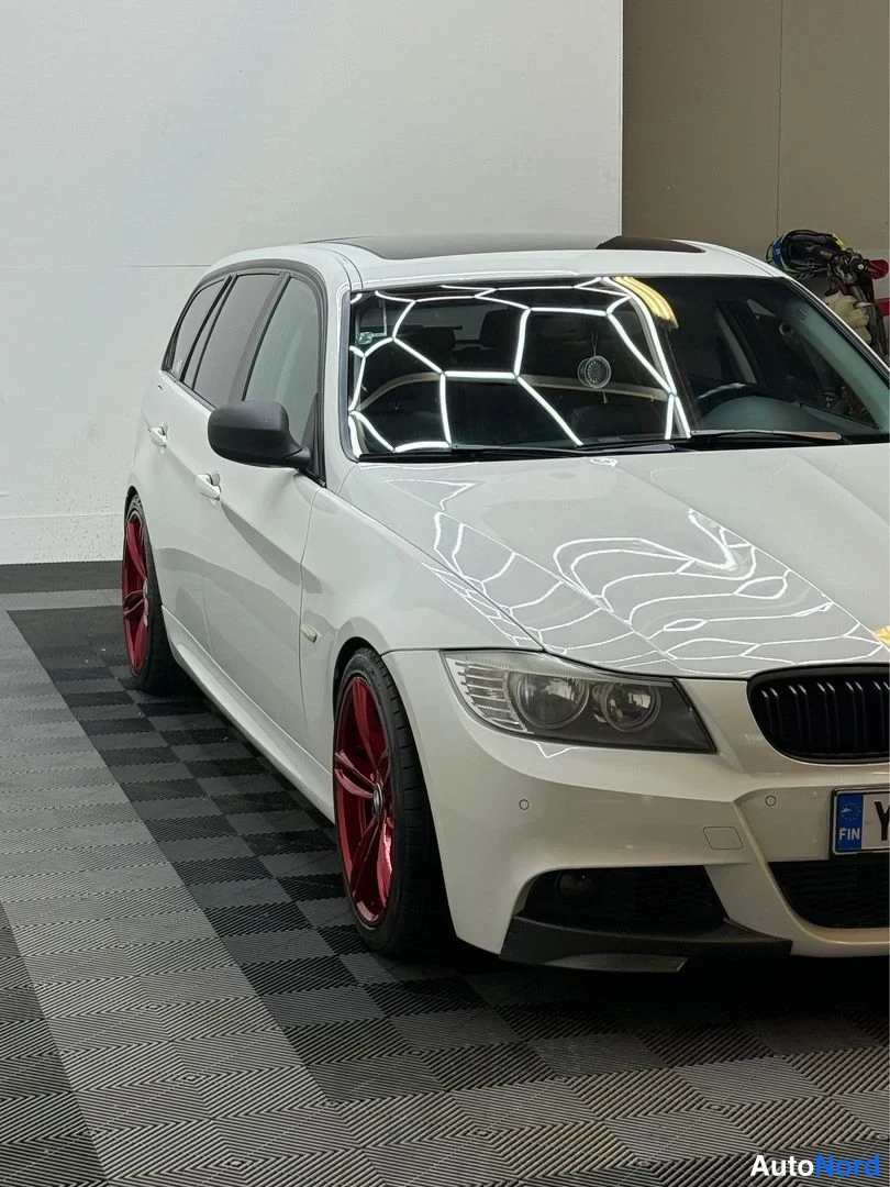 Etuviisto, nokka oikealle: Valkoinen BMW-farmari etukolmasosa oikealla, punaiset vanteet, sisätiloissa.