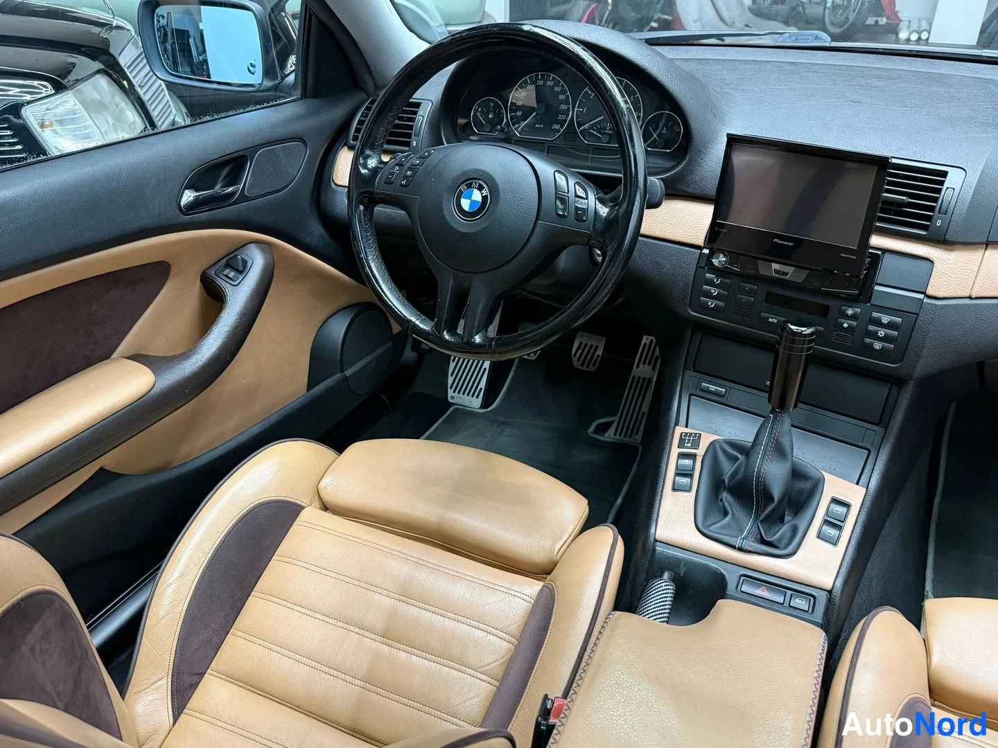 Edestä: Auton sisätilat kuljettajan oven kohdalta kuvattuna: beige-nahkainen istuin, BMW-ohjauspyörä, Pioneer-näyttö ja vaihtokeppi.