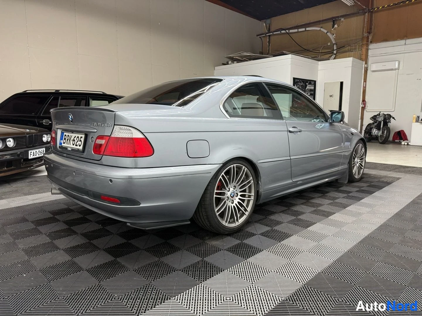 Takaviisto: Hopeanharmaa BMW-coupé takaosaa ja oikeaa kylkeä hallissa; rekisteritunnus, sauteri keulan logo.