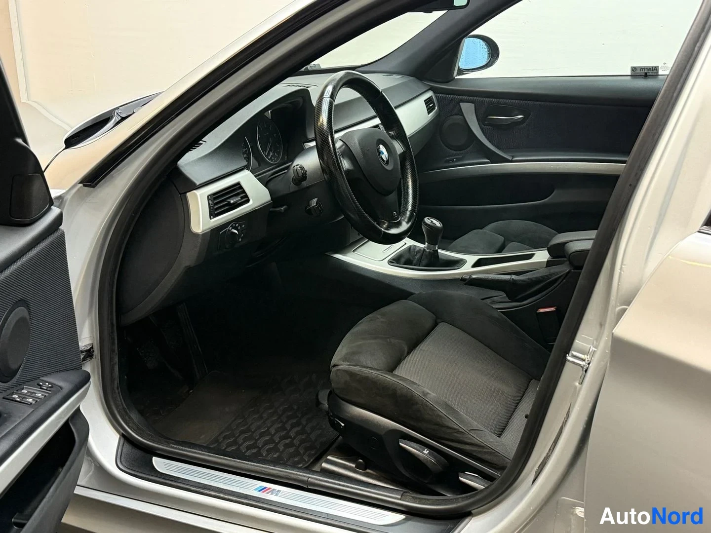Muu: Sisätiloista otettu kuva, joka näyttää BMW:n ohjaamon vasemman sivun: koristeellinen korimoustoiminen ohjauspyörä, manuaalivaihdevipu ja M-kylttikynnyslista.