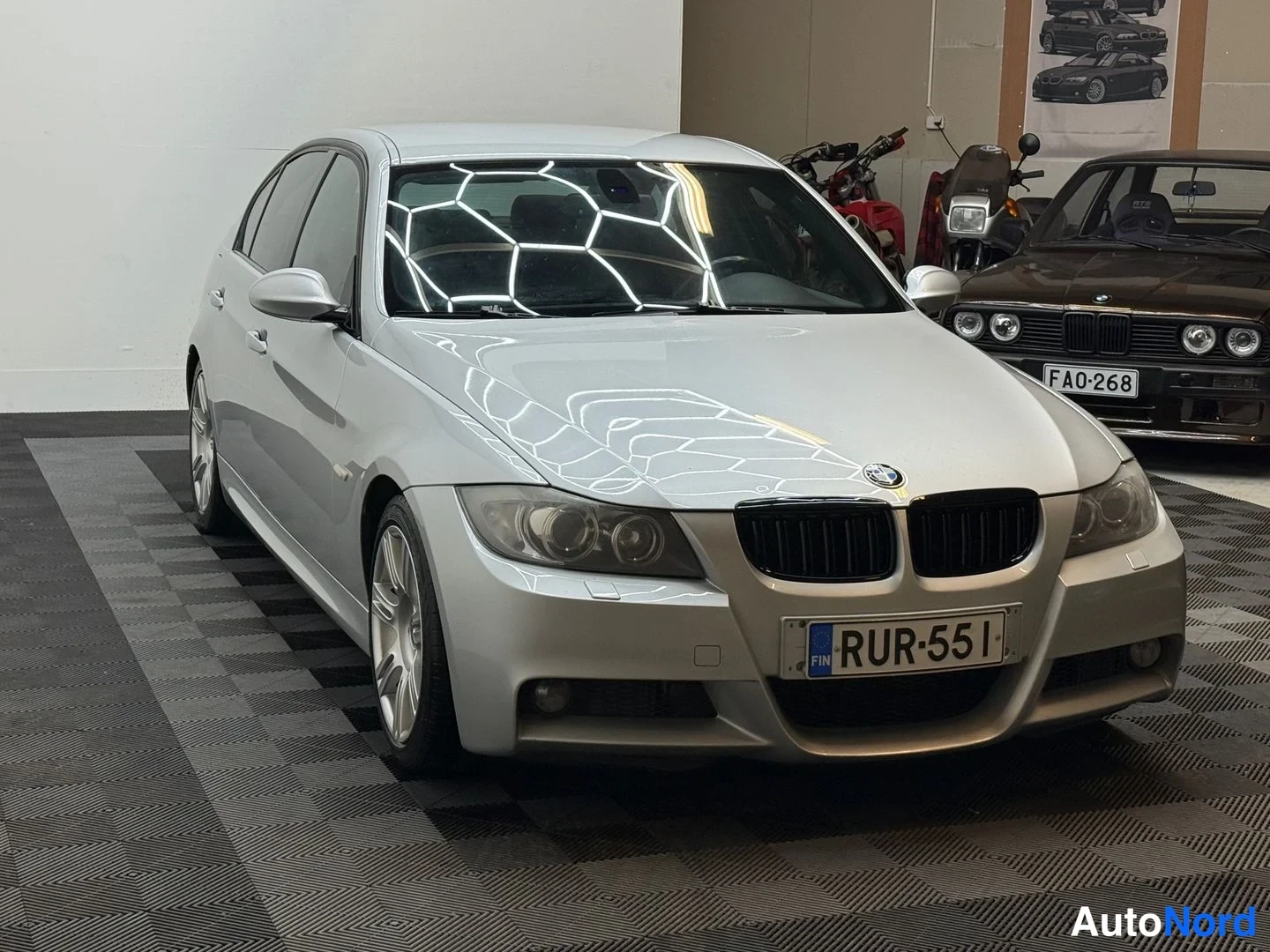 Etuviisto, nokka oikealle: Hopea BMW-sedan edessä-vino oikealla kuvakulmassa, taustalla toinen auto ja moottoripyöriä.