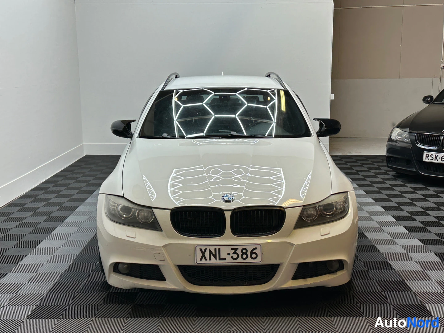 Edestä: Valkoisen BMW:n suora etunäkymä, rekisterikilpi, etuspoileri ja sumuvalot näkyvissä; oikealla kulmassa toinen auto.