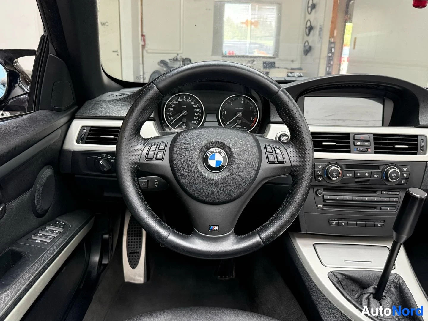 Sivulta: Näkymä ohjauspyörään ja kojelautaan: BMW-logollinen M-ohjauspyörä, mittaristot, ikkunakytkimet ovessa ja keskikäyttöpöydän radio- ja ilmastointisäätimet.