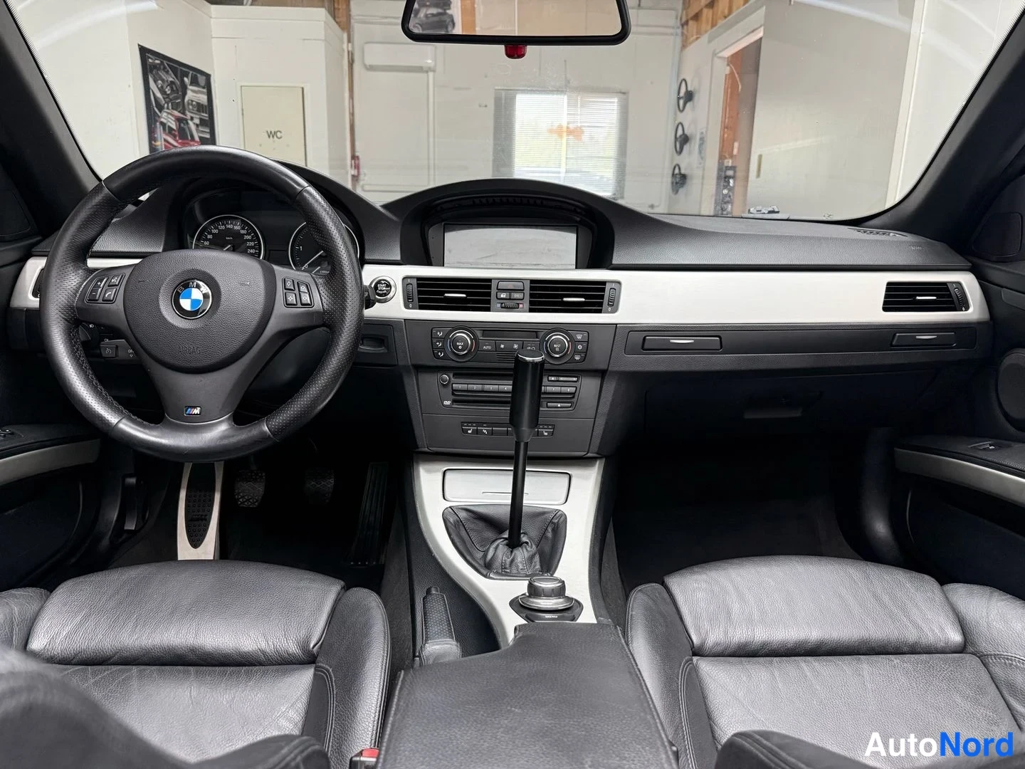 Sivulta: Etusisätila: BMW-ohjauspyörä ja analogiset mittarit, keskikäyttöpöydän näyttö, manuaalivaihtajan kehys, mustat nahkaistuimet ja harmaat koristelistat.