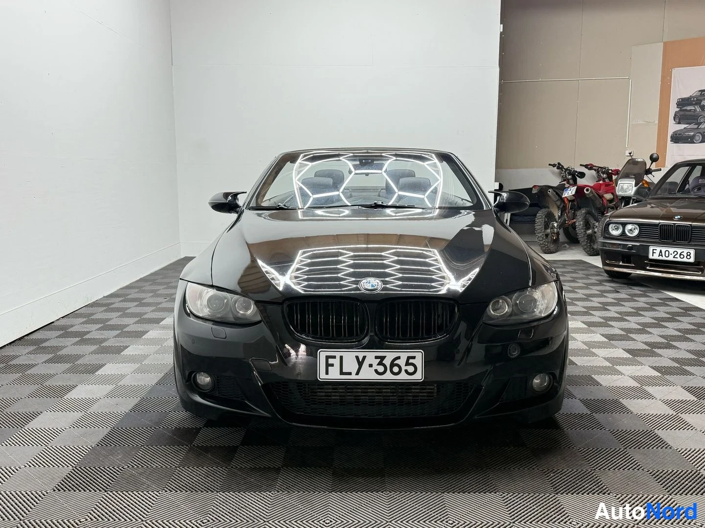 Edestä: Mustan BMW:n suora etunäkymä, rekisteritunnus, sumuvalot ja sumuvalot, taustalla moottoripyöriä ja toinen auto.