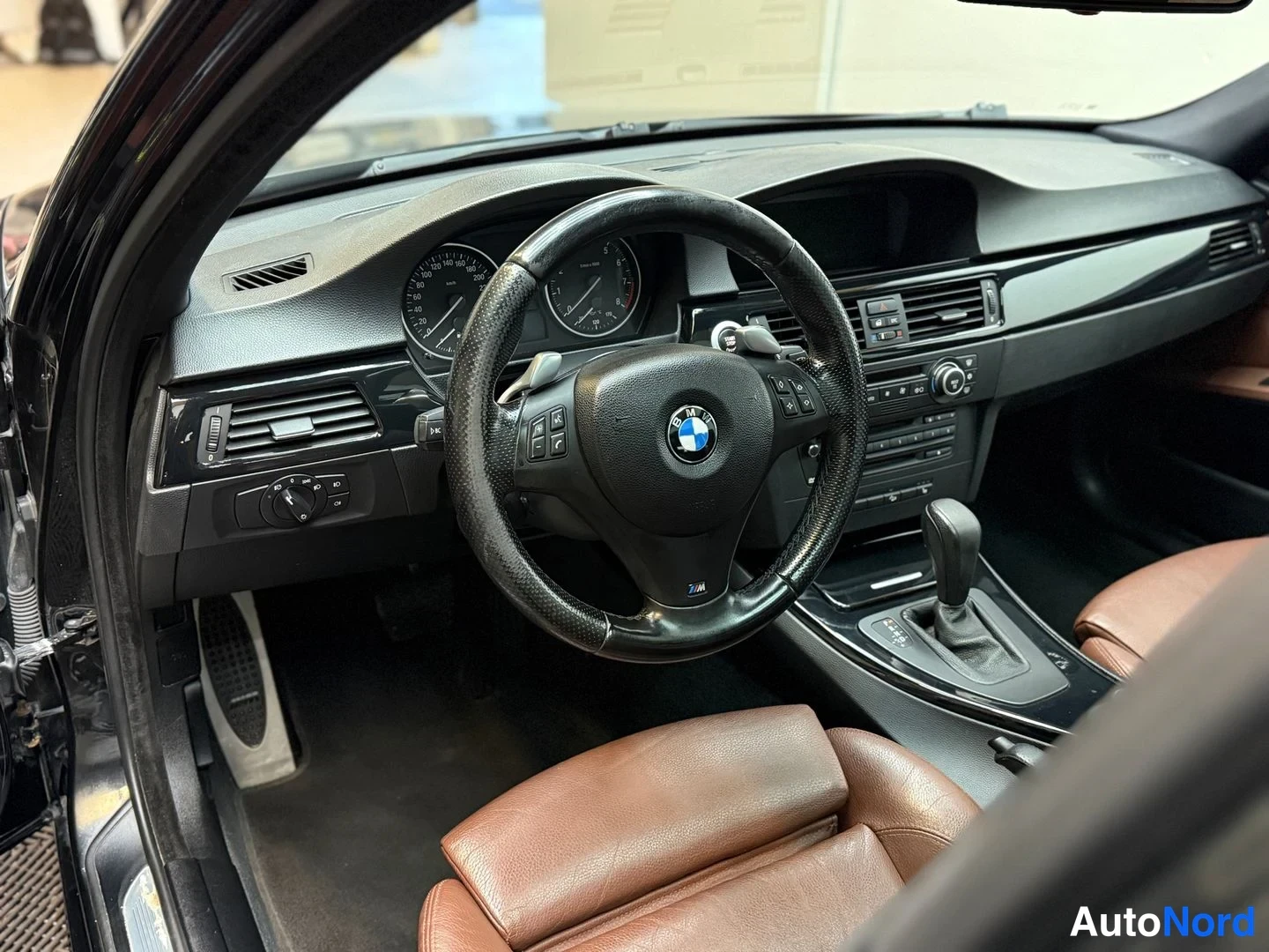 Muu: Ohjauspyörä ja kojelauta lähikuvassa: BMW-merkki, mittarit, automaattivaihteen valitsin ja ruskeat nahkaistuimet.