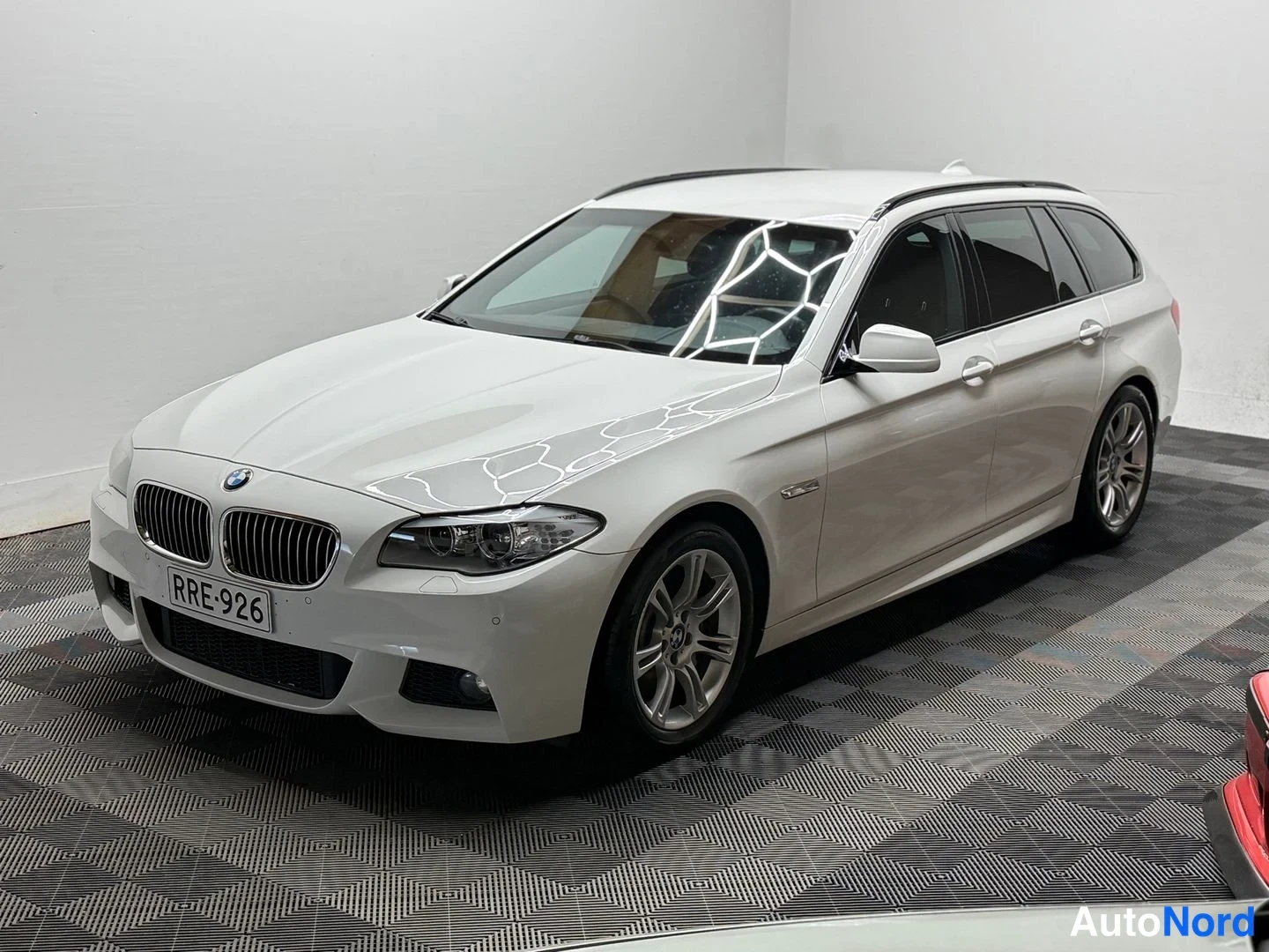 Etuviisto, nokka vasemmalle: BMW 525d -farmariauto kolmasosa edestä vasemmalta, valkoinen korimaali, rekisterikilpi, sisätilassa harmaata lattiaratkaisua vasten.