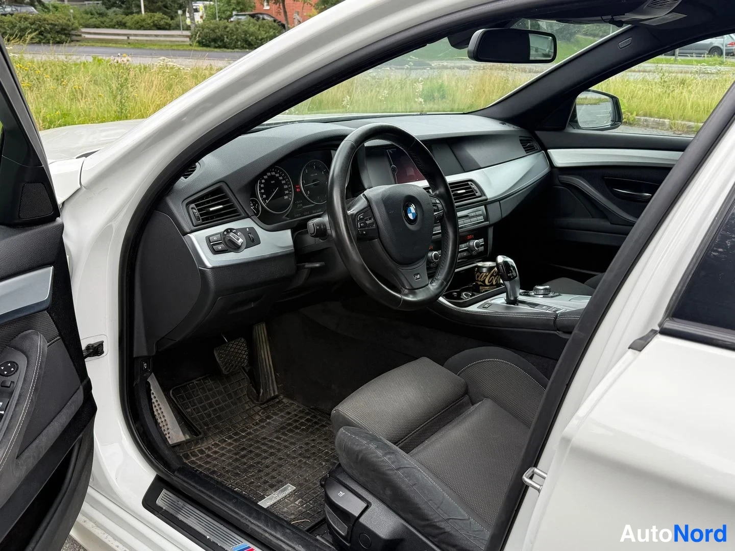 Muu: Auton etuosa sisätilat: BMW-ohjauspyörä ja mittaristo, musta sisusta, kuljettajan ovi auki.