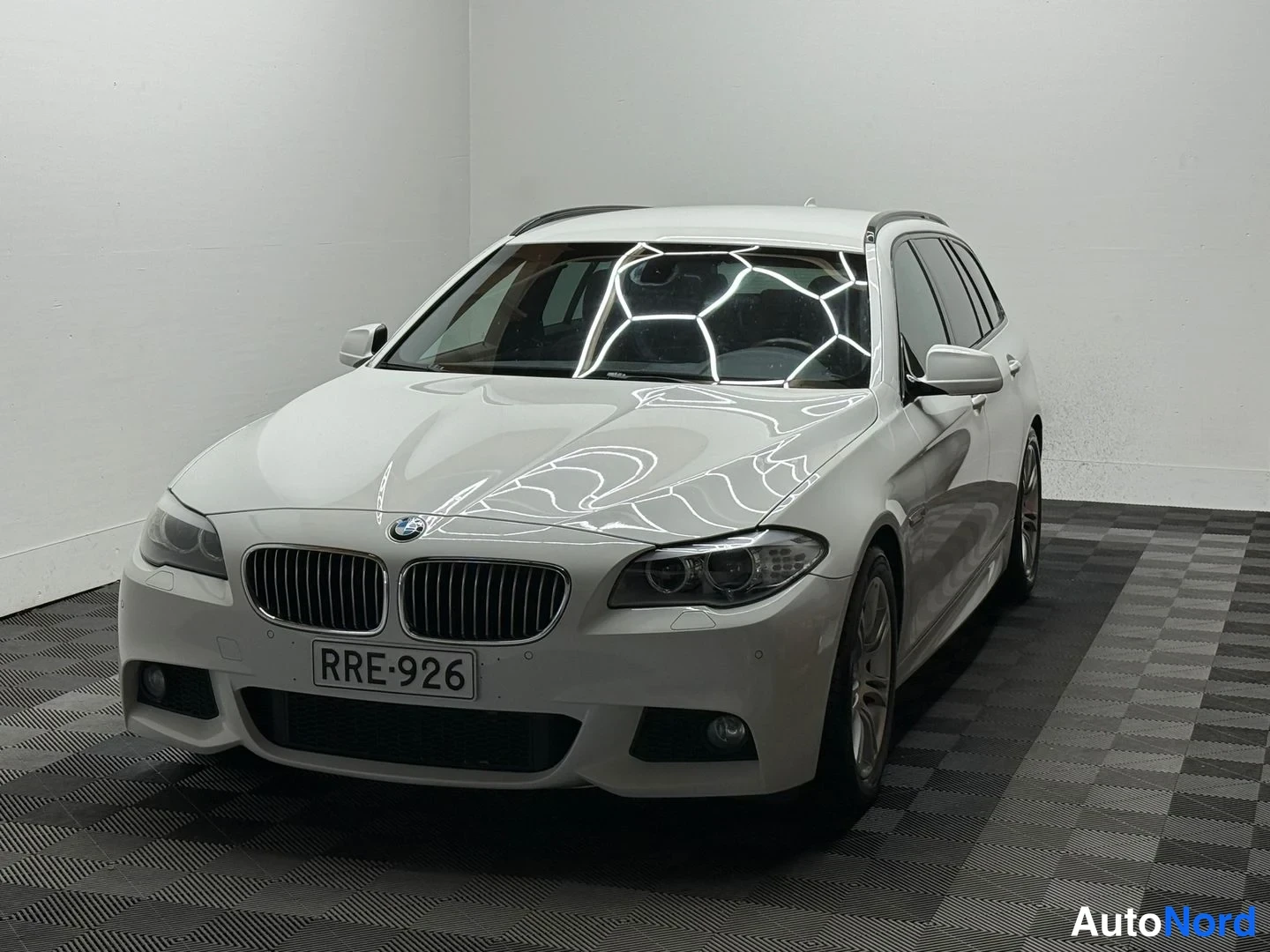 Etuviisto, nokka vasemmalle: BMW 525d -farmariauto etukulmasta, valkoinen korimaali, rekisterikilpi, BMW-merkki konepellissä, sisätilassa valkoista seinää vasten.