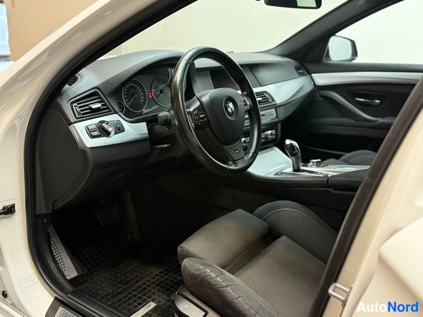 Edestä: Sisäkuva avoimesta kuljettajan ovesta: BMW-ohjauspyörä, mittaristo, automaattivaihteen valitsin ja tummat istuimet.