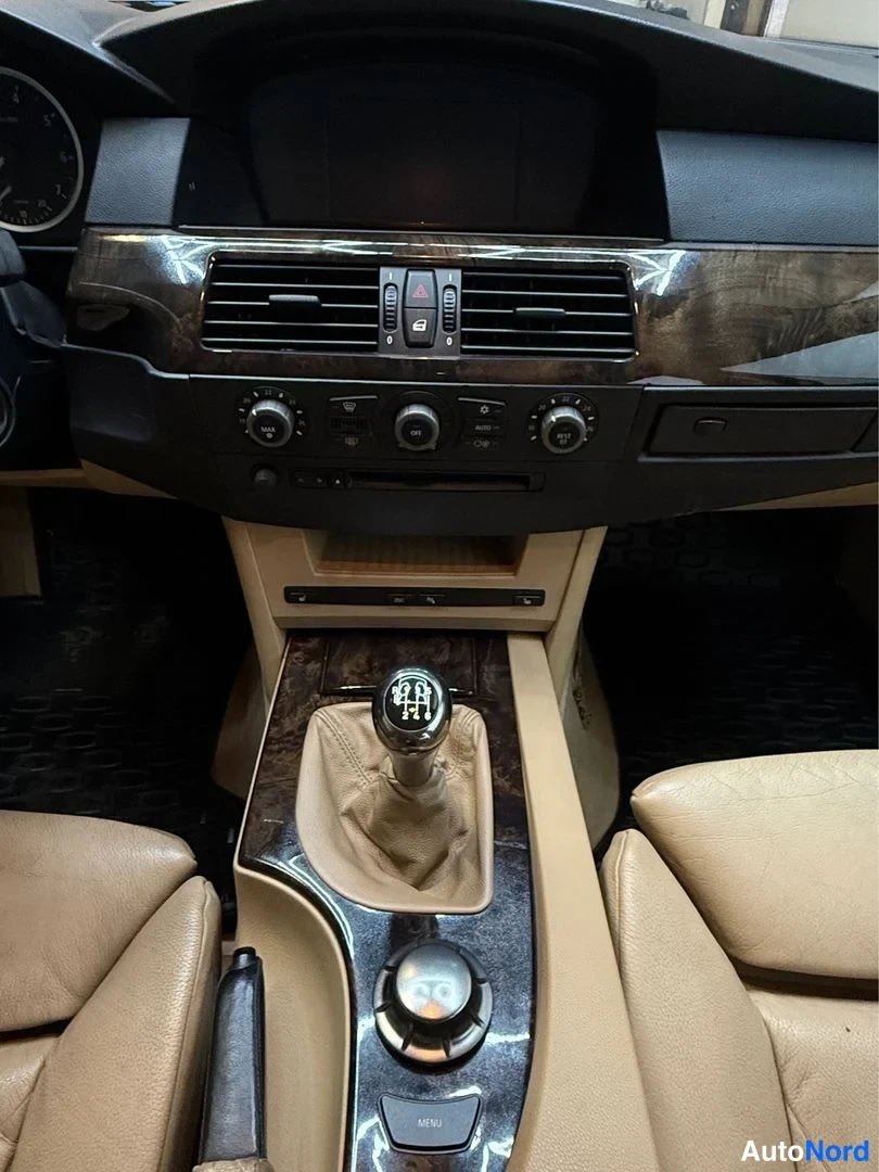Muu: Auton kojelaudan ja keskikonsolin lähikuva: manuaalivaihteerin vipu, iDrive-ohjain ja ilmastointilaitteen säätöt, beige nahkapinnat.