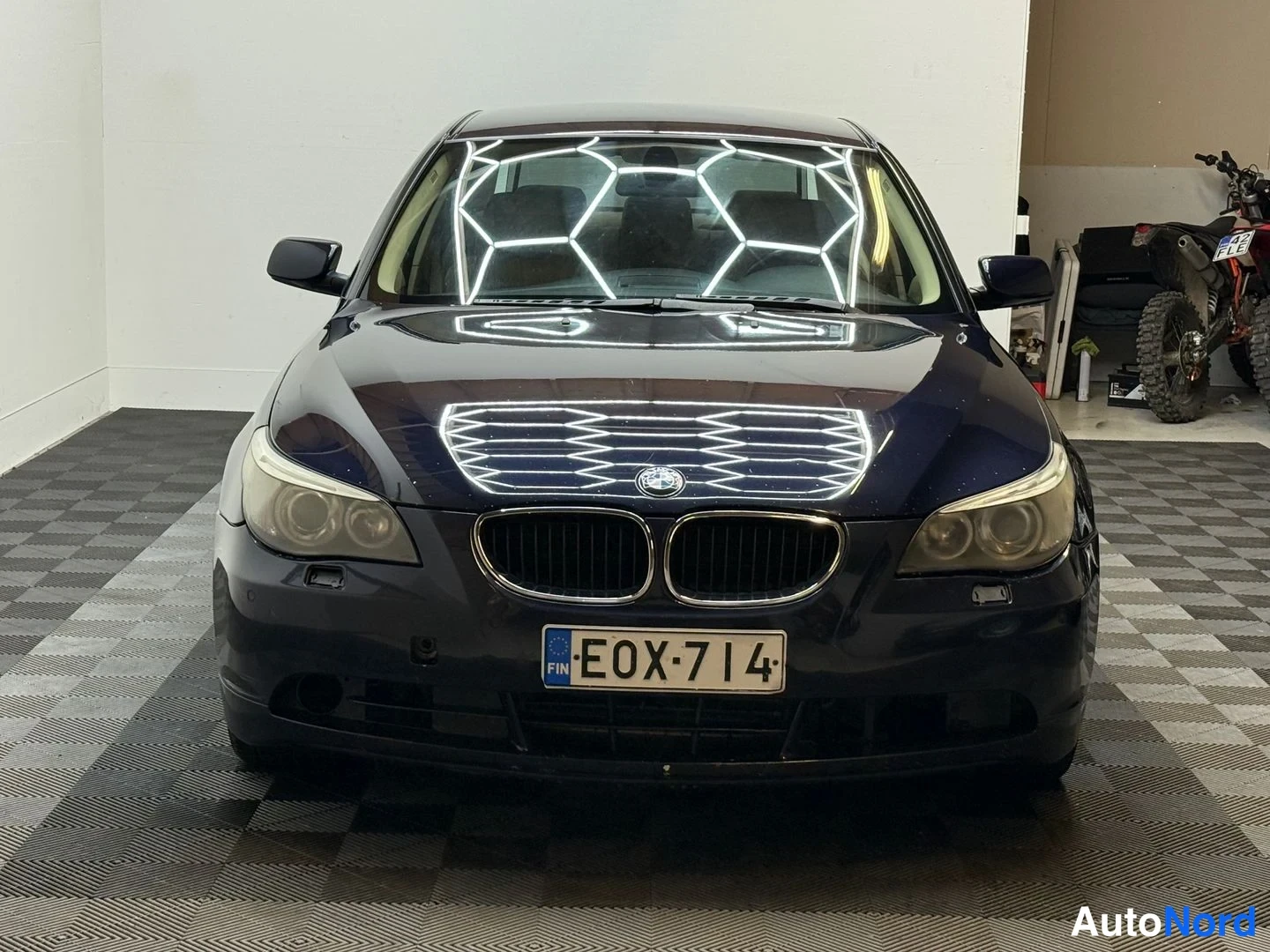 Edestä: Sininen BMW-sedan suoraan edestäpäin kuvattuna, rekisterikilpi, huone valkoinen seinä ja ruutumatto lattialla, taustalla moottoripyörä.