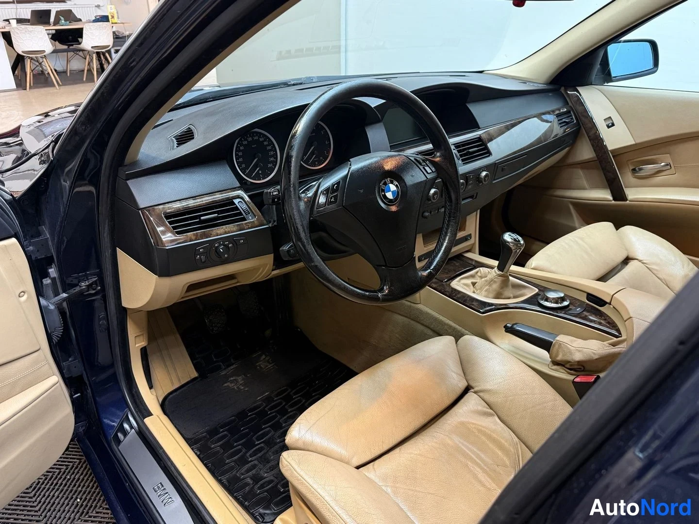 Sivulta: Näkymä auton sisätiloihin kuljettajan oven auki: BMW-merkkinen rattin, mittaristo, käsivaihteet ja beige nahkapenkit.