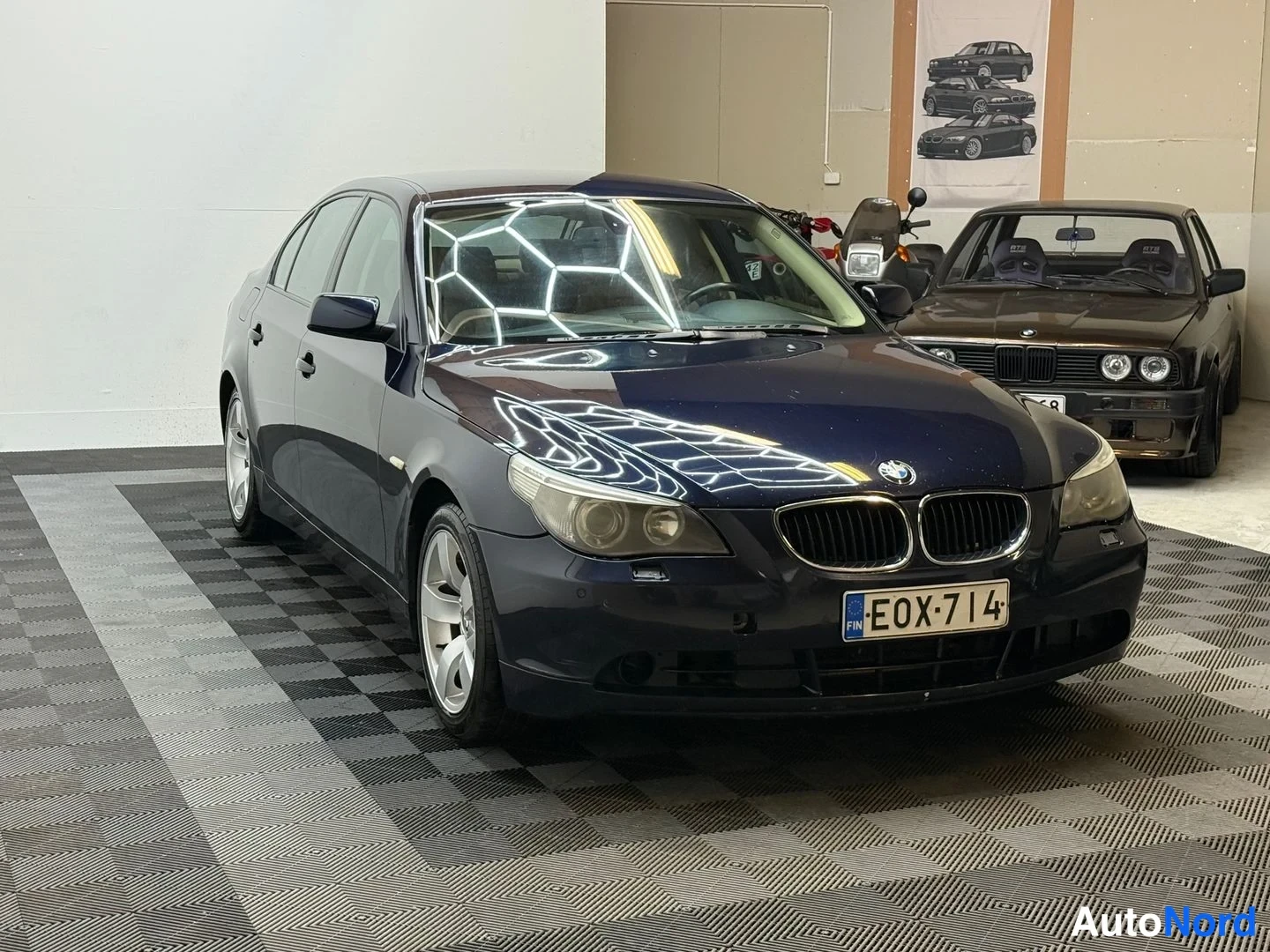 Etuviisto, nokka oikealle: Sininen BMW-sedan kulmassa edestä oikealta kuvattuna, rekisterikilpi, taustalla vanhempi BMW-auto ja seinällä juliste.