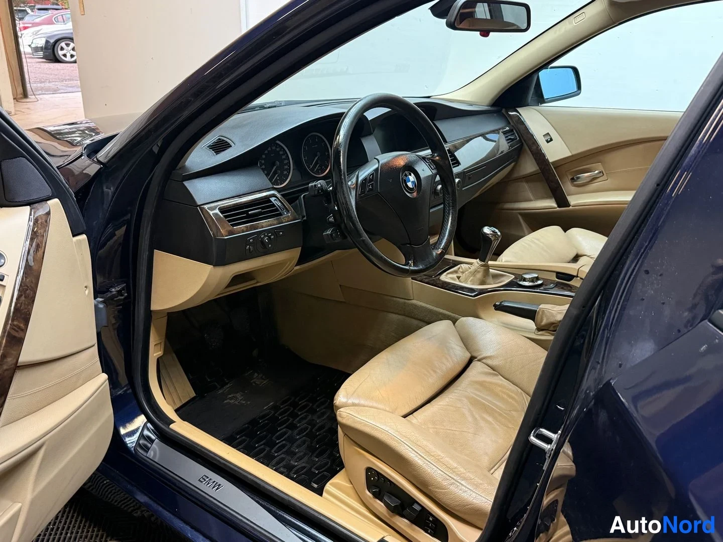 Sivulta: Auton sisätilat kuljettajan puolelta: BMW-rattin, mittaristo, vaihteen­keppi ja beige nahkapenkit, ovi auki.