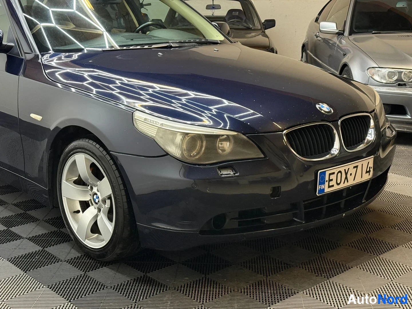 Etuviisto, nokka oikealle: Auton etukulma: tummansininen BMW, munuaiskehys, ajovalot ja -etukilpi näkyvät, kevytmetallivanne etupyörässä.