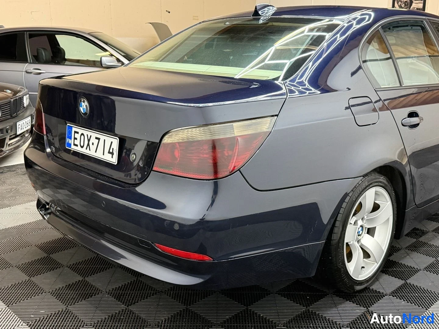 Takaviisto: Auton takakulma: tummansininen BMW-sedan, punaiset takavalot, kannen BMW-tunnus ja E0X-714-rekisterikilpi näkyvät.
