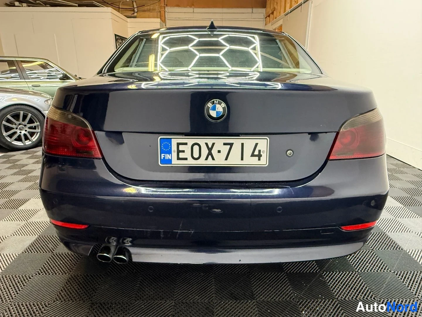 Takaa: Sinertävän tumma BMW-sedanin perä suoraan takaa; rekisterikilpi, BMW-embleemi, punaiset takavalot ja kaksi pakoputkea vasemmalla.