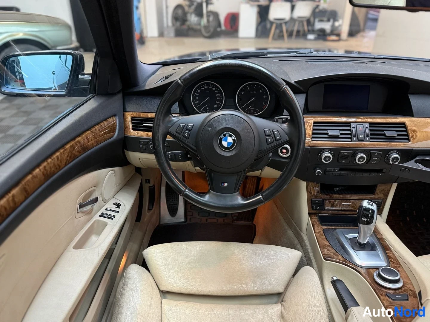 Etuviisto, nokka vasemmalle: Näkymä ohjaamoon ajoneuvon ulkopuolelta: BMW-ohjauspyörä, mittarit, puukuviollinen kojelauta, automaattivaihteiston valitsin ja beige-istuin.