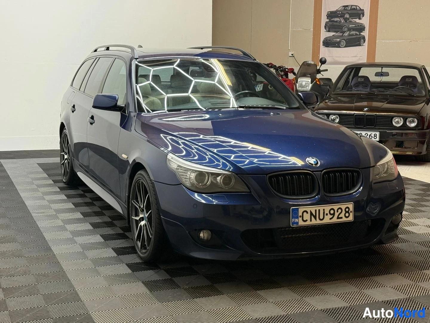Etuviisto, nokka vasemmalle: Tummansininen BMW-farmari keulakulmasta toisesta suunnasta, kiiltävä korimaali heijastaa valoja, rekisterikilpi.
