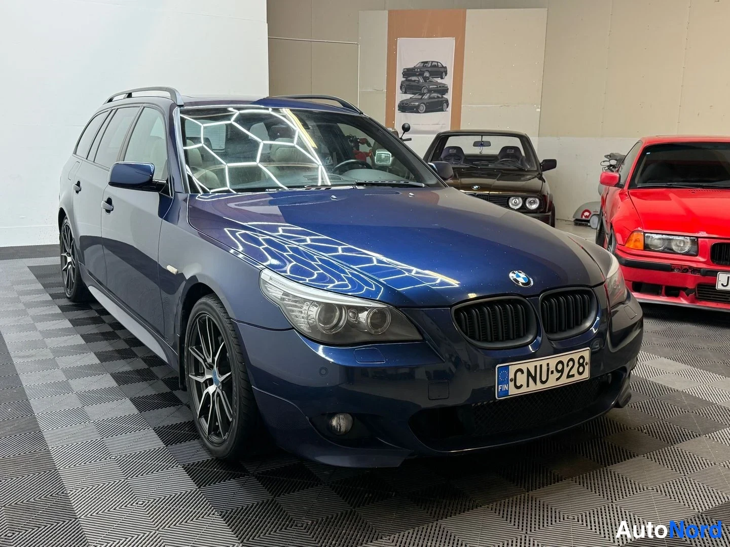 Etuviisto, nokka oikealle: Tummansininen BMW-maasturi edessä sivulta oikeaan nojaavassa kulmassa sisätilassa, rekisterikilpi.