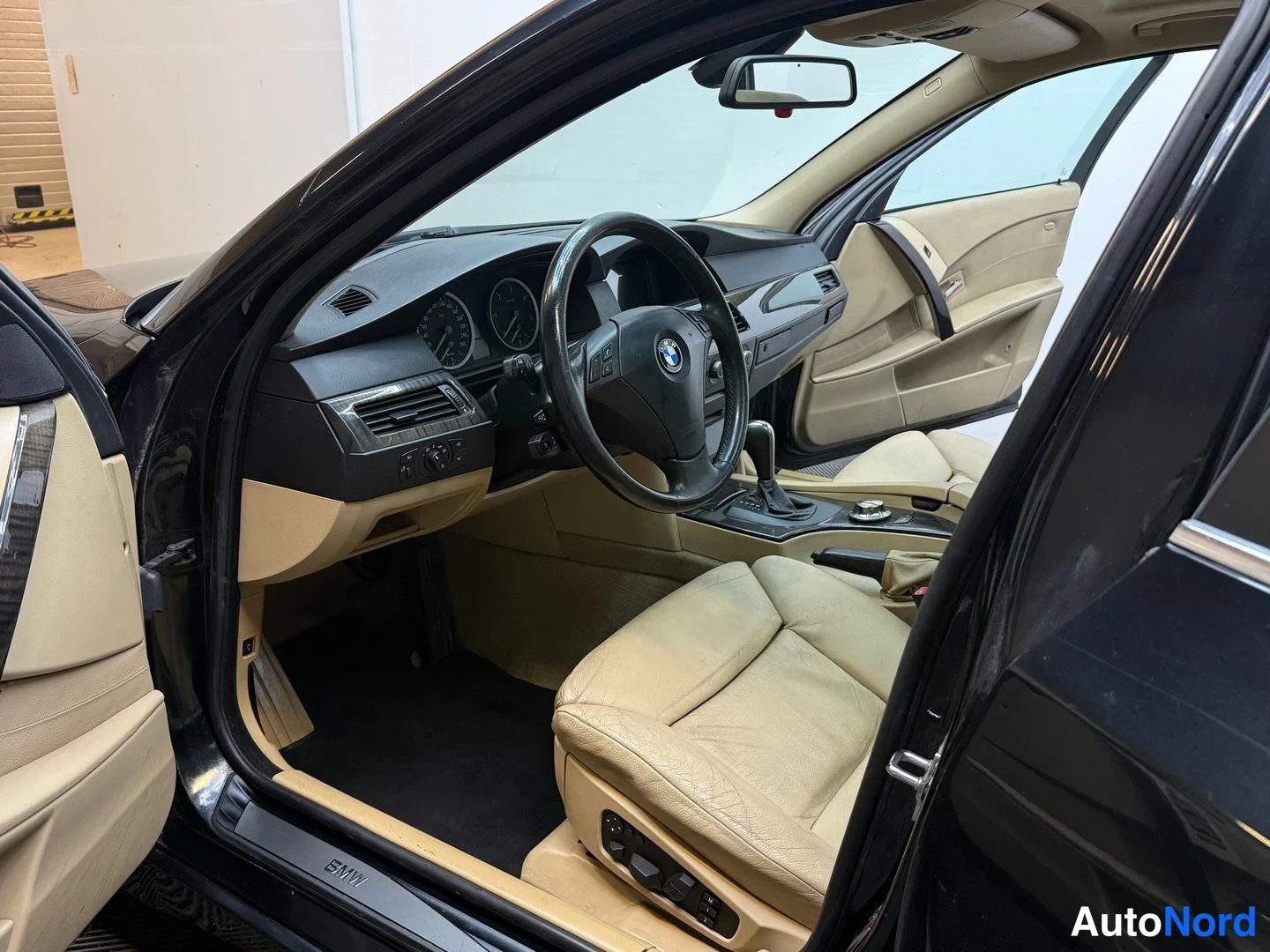 Muu: Avoin kuljettajan ovi, ohjaamo jossa beige nahkapenkit, BMW-ohjauspyörä ja tumma kojelauta.