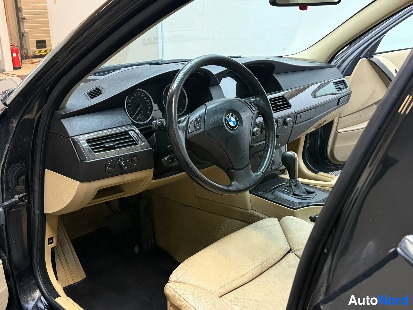 Muu: Ohjaamon näkymä: BMW-merkillinen ohjauspyörä, analogiset mittarit, tumma kojelauta ja beige istuimet.