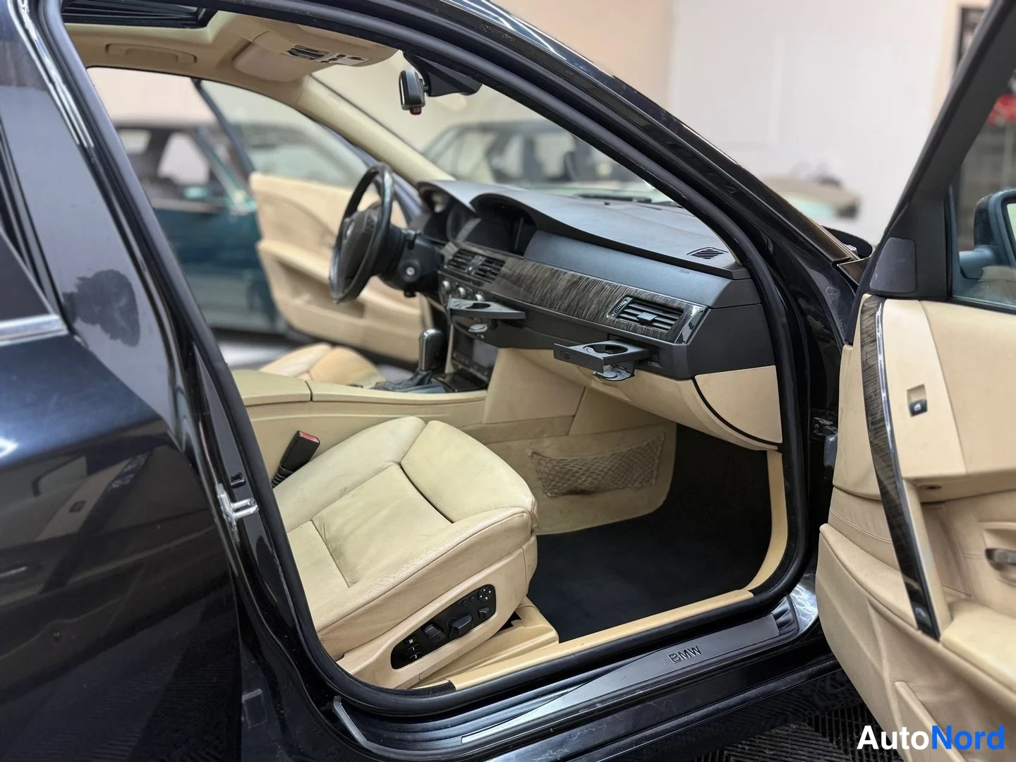 Muu: Auton sisätilat: avoimen kuljettajan oven takaa näkyy beige nahkapenkit, tumma kojelauta, ohjauspyörä ja BMW-merkillinen kynnyslista.