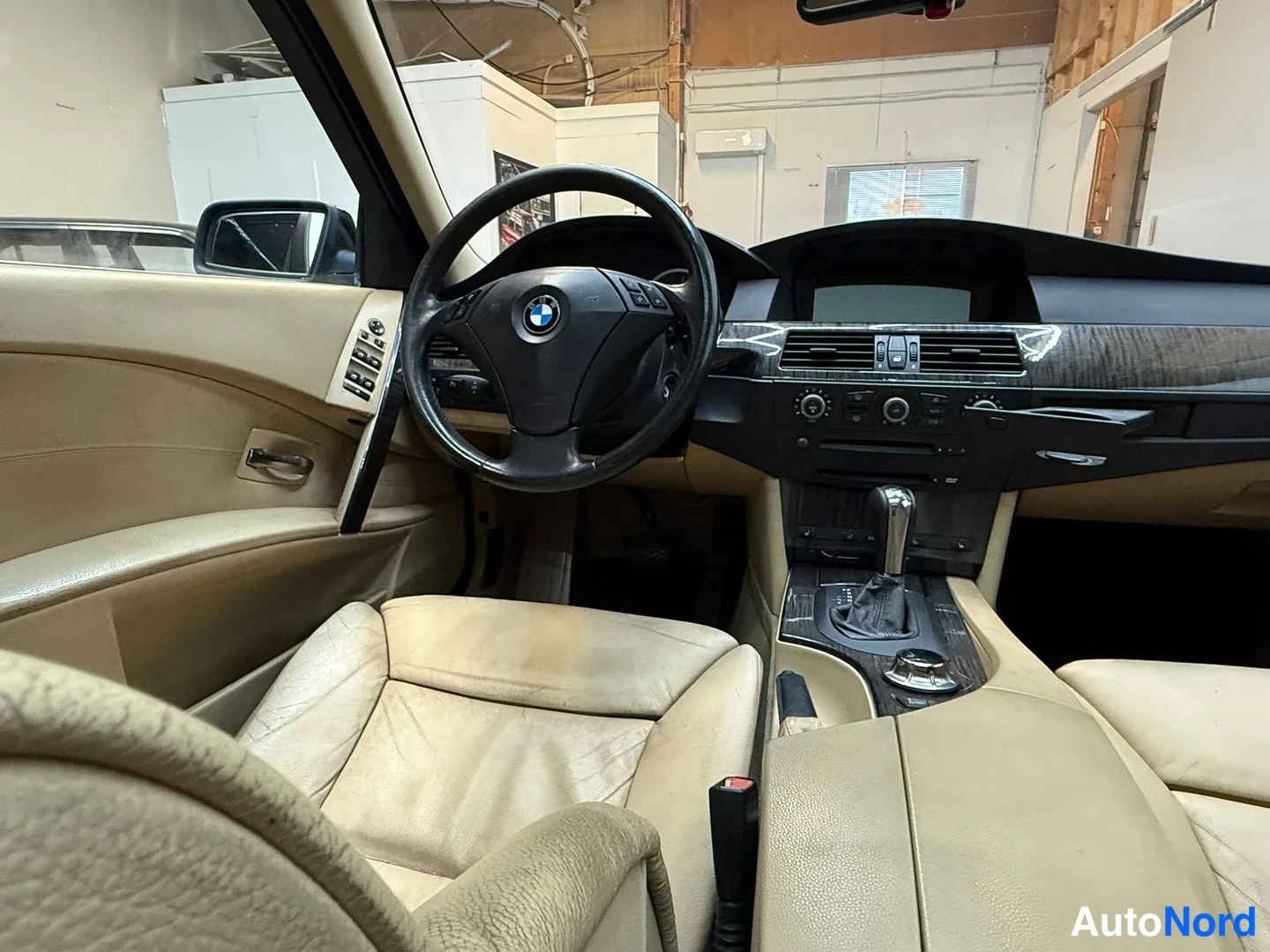 Muu: Kuljettajan puoleinen näkymä BMW:n ohjaamoon: BMW-merkkinen ohjauspyörä, tumma kojelauta, aurtomaattivaihteen vaihtajakatkaisin ja beigeä nahkaverhoilu.