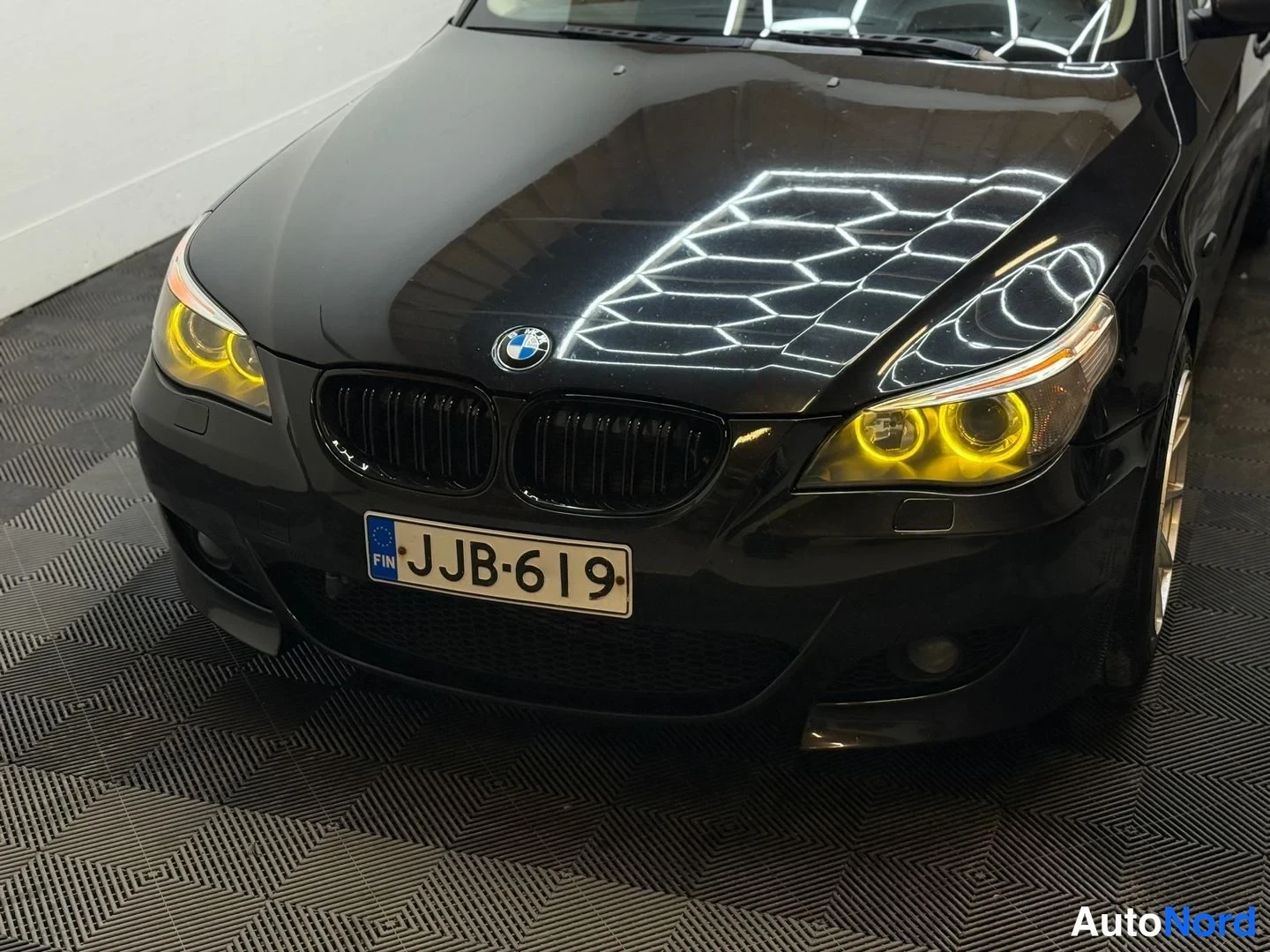 Edestä: Auton etupäätä suoraan edestä: musta BMW-kori, -rekisterikilpi ja keltarenkaiset etuprojektörit.