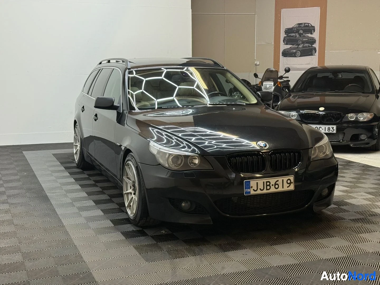 Etuviisto, nokka oikealle: Musta farmariauto (BMW) nähtynä etu-oikealta kolmasosalta, ET-kilpi, kattotankokaaret ja metallin väriset kevytmetallivanteet näkyvissä.