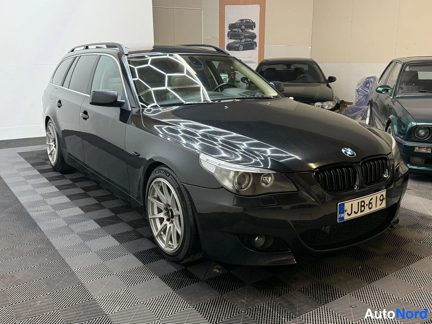 Etuviisto, nokka oikealle: Musta farmariauto (BMW) nähtynä etu-oikealta kolmasosalta, ET-kilpi, kattotankokaaret ja metallin väriset kevytmetallivanteet näkyvissä.