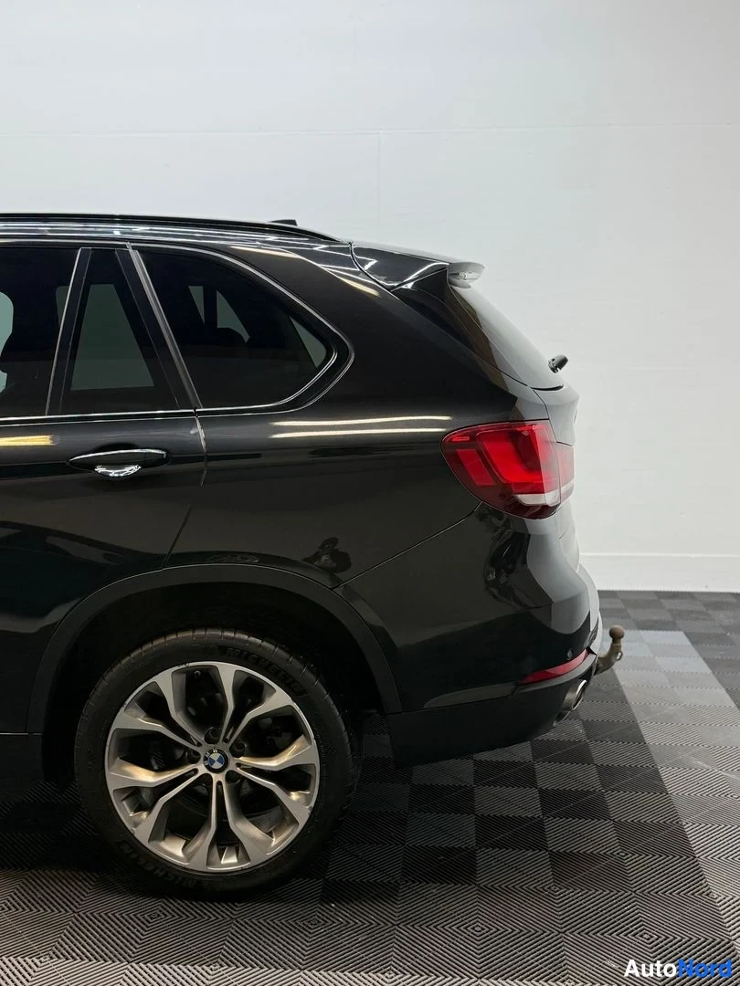 Takaviisto: BMW X5:n takakolmasosakuva: punainen takavalo, musta kori ja kevytmetallivanne hallin harmaassa lattiassa.