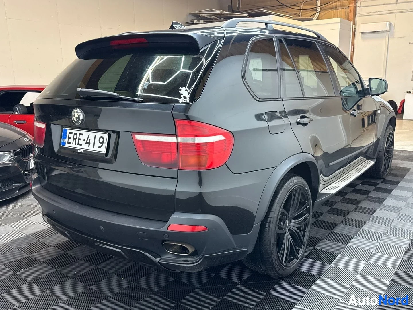 Takaviisto: Mustan SUV-auton takaosa ja oikea kylki hallitilassa; rekisterikilpi, kattokaiteet, askelmat ja mustat vanteet näkyvissä.