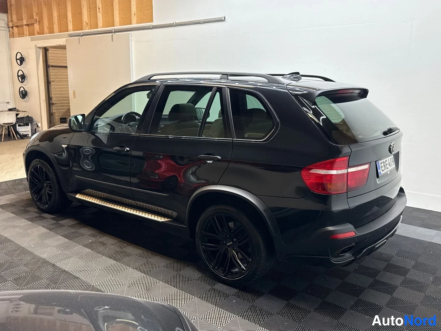 Takaviisto: BMW X5:n takaosa ja vasen kylki sisätiloissa, musta maali, kattotankotangot ja mustat vanteet näkyvissä.