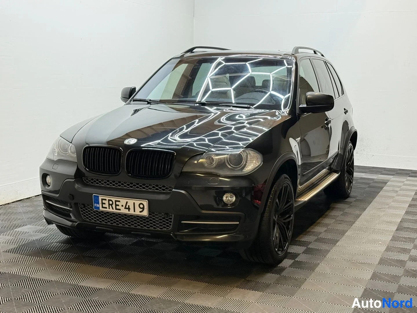 Etuviisto, nokka vasemmalle: Musta BMW X5 etuviistosta vasemmalle, kilpi, sivulaskuimet ja mustat vanteet.