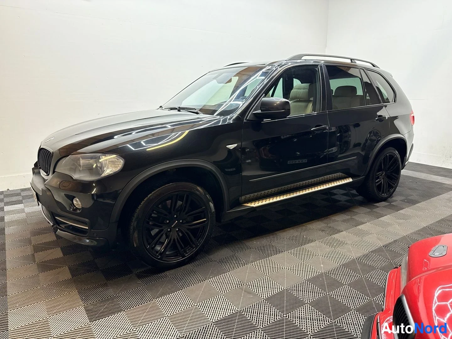 Etuviisto, nokka vasemmalle: Mustan BMW X5:n etukulmanäkymä sisätiloissa; näkyvissä kattokaiteet, sivulauttarit ja mustat kevytmetallivanteet.