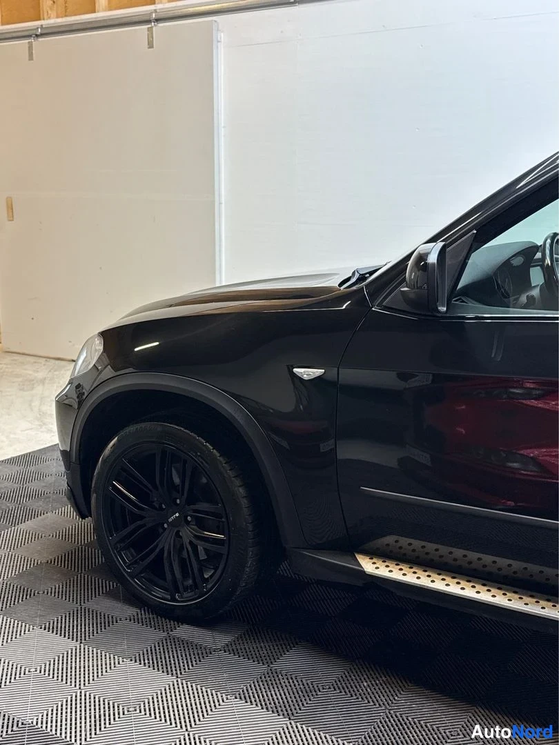 Etuviisto, nokka vasemmalle: Mustan BMW X5:n etupään sivunäkymä garage-tilassa; näkyvissä etupyörä mustalla vanteella ja sivulauttari.