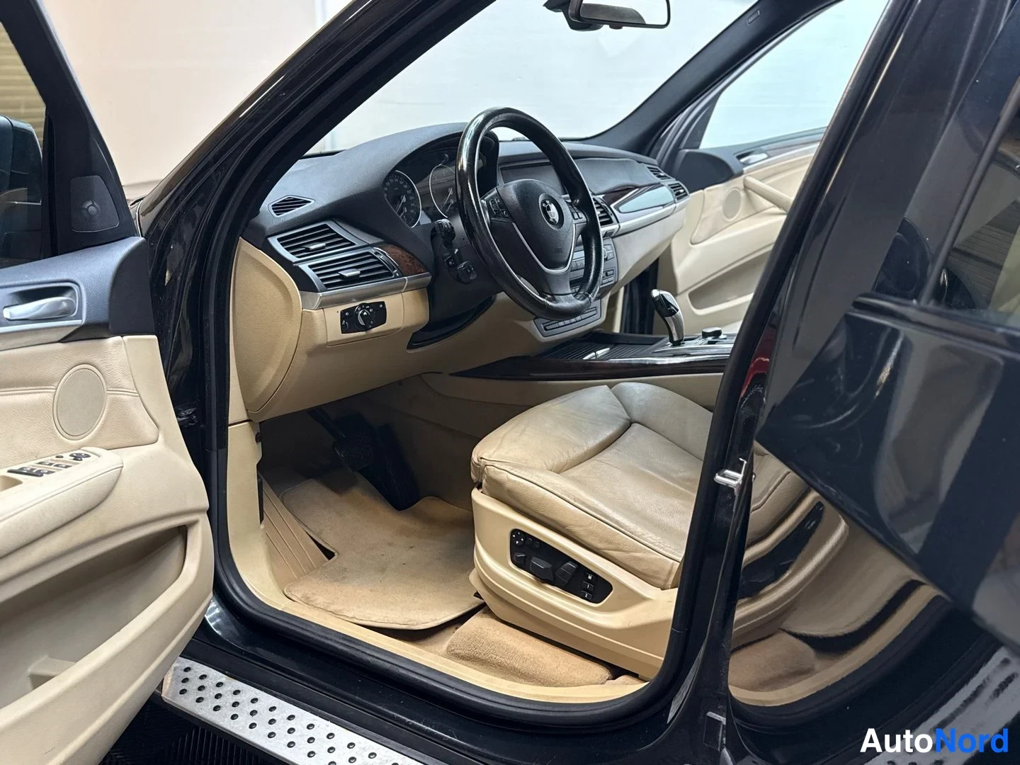 Muu: Kuljettajan puolen ovi auki: beige sisusta, BMW-ohjauspyörä, tummat ovipaneelit ja metallinen kynnyslista näkyvät.