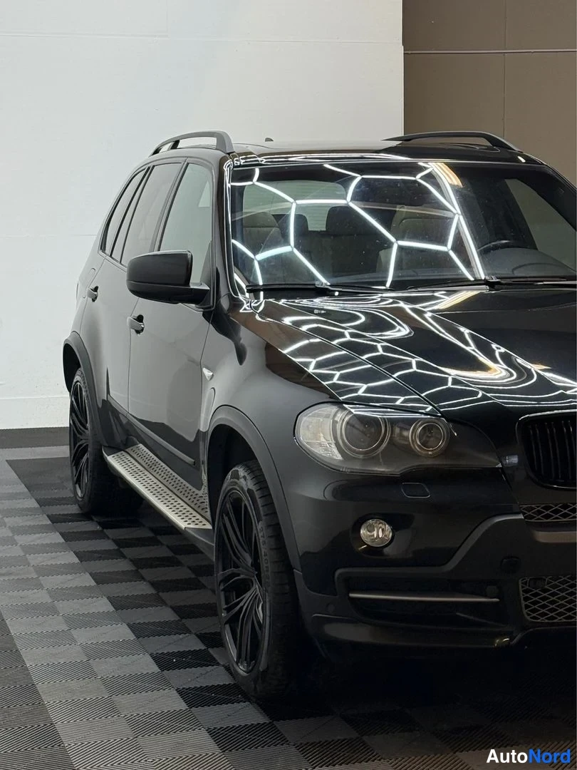Etuviisto, nokka oikealle: Mustan BMW X5:n etukulma sisätiloissa; etupuskuri, ajovalot ja kylki näkyvät, takapää on kuvan ulkopuolella.