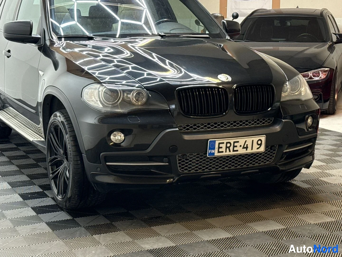 Etuviisto, nokka oikealle: Mustan BMW X5:n etukulma; etupanssari, etusuoja- ja ajovalot, kylkiaskelmat ja mustat vanteet näkyvissä; rekisteritunnus.