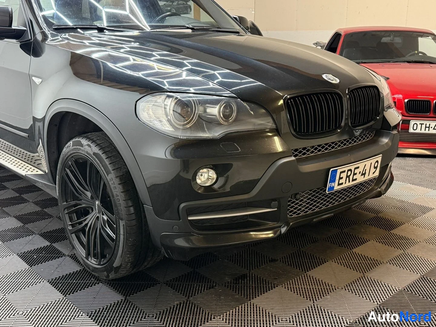 Etuviisto, nokka oikealle: BMW X5:n etukulma mustana, rekisteri, mustat vanteet, tuulilasi ja konepelti näkyvät sisätiloissa.