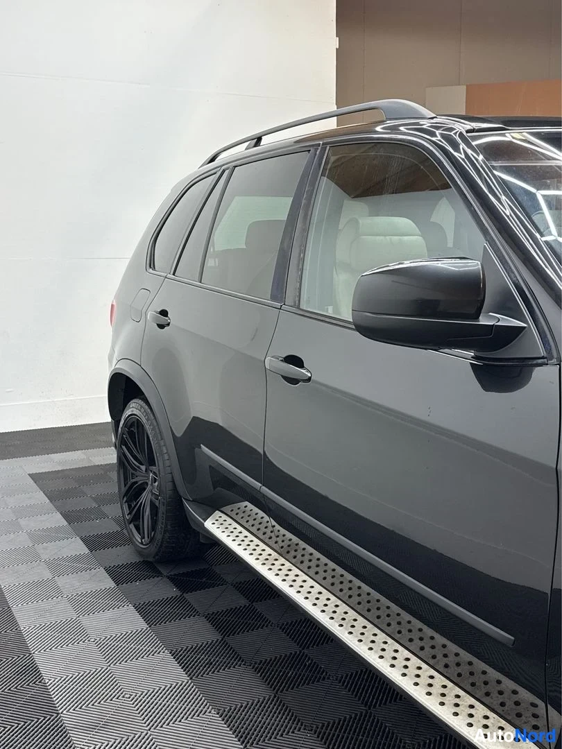 Sivulta: Mustan SUV-auton oikea sivuprofiili sisätiloissa; näkyvissä kattokaiteet, ovien kahvat, mustat kiiltelevät vanteet ja metalliset askelmat.