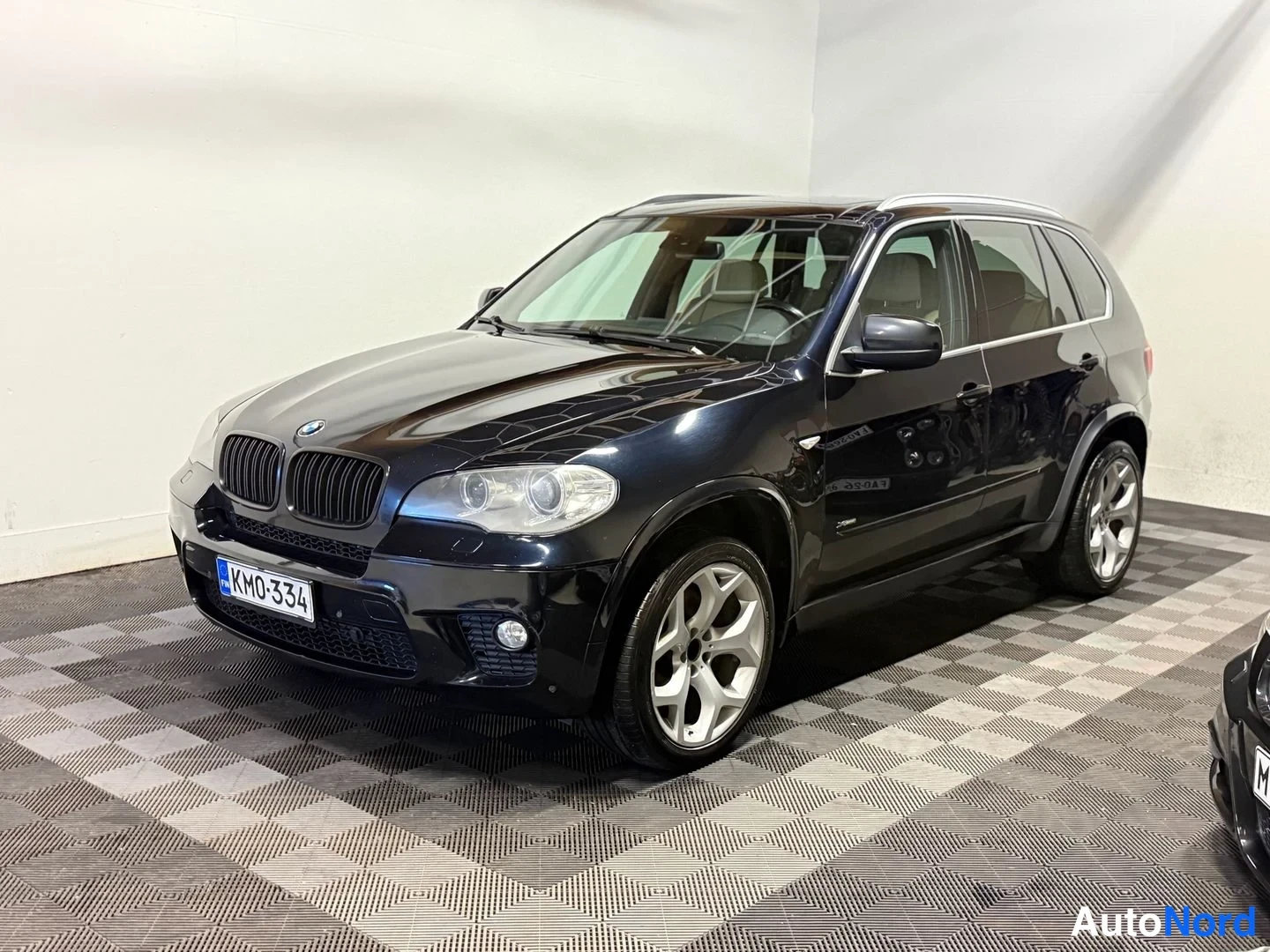 Etuviisto, nokka vasemmalle: Musta BMW X5 -maastauto kolmannelle neljännekseltä edestä vasemmalla sisätiloissa ruudullisella lattialla, rekisterikilpi.