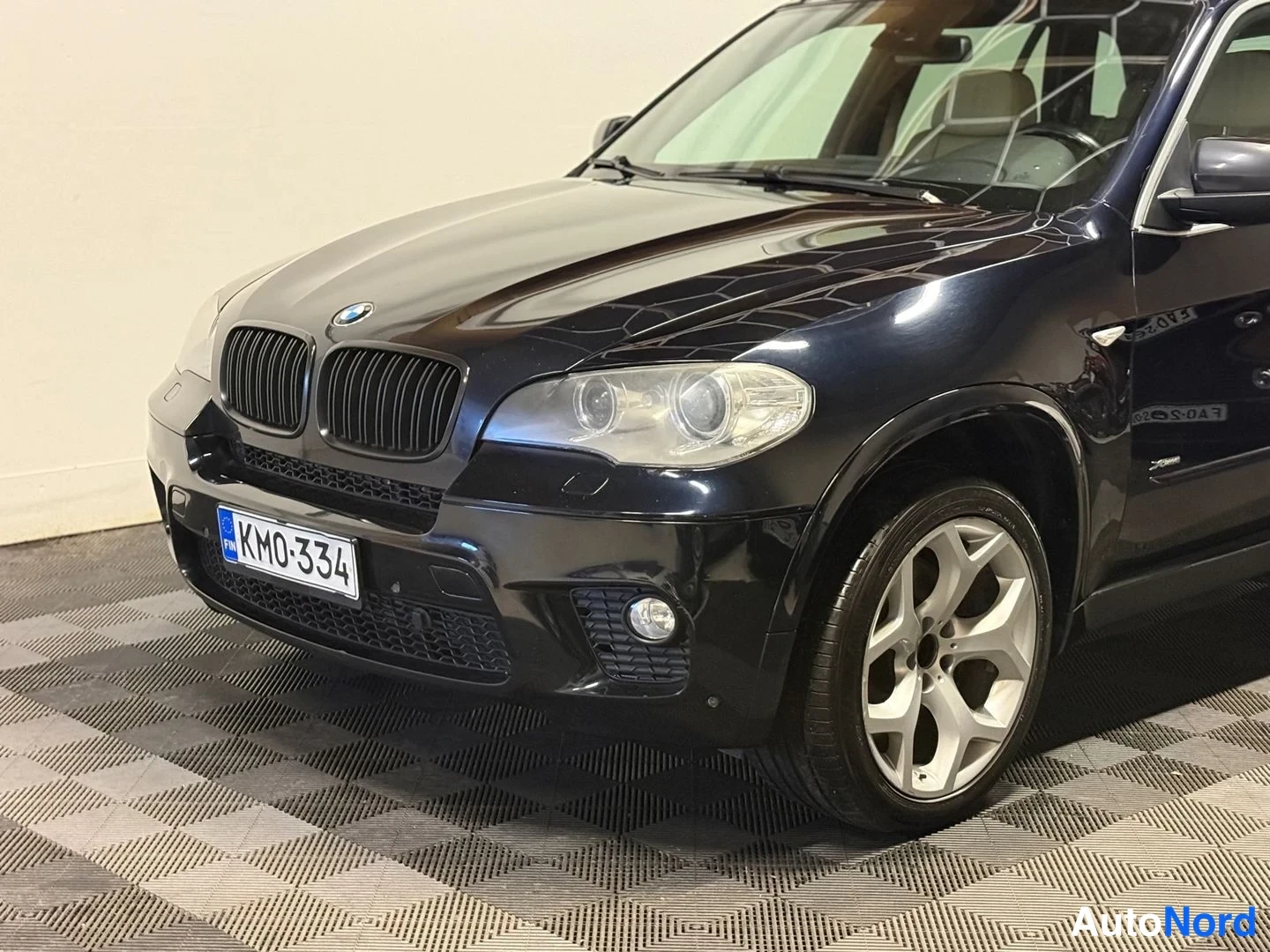 Etuviisto, nokka vasemmalle: Lähikuva mustan BMW X5:n keulasta vasemmalta, etusäleikkö, ajovalot ja rekisterikilpi näkyvissä.