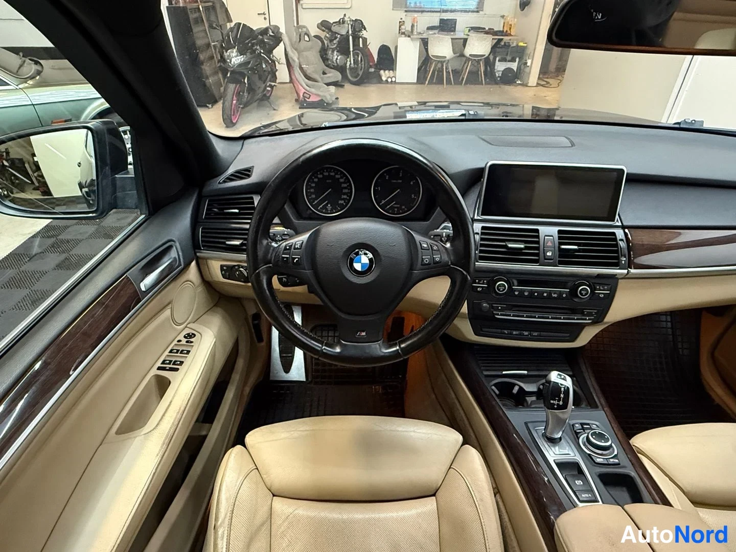 Muu: Auton sisätila ohjaamosta kuvattuna: BMW-merkkinen ohjauspyörä, mittarit, keskikonsolin näyttö ja vaaleat nahkaistuimet.