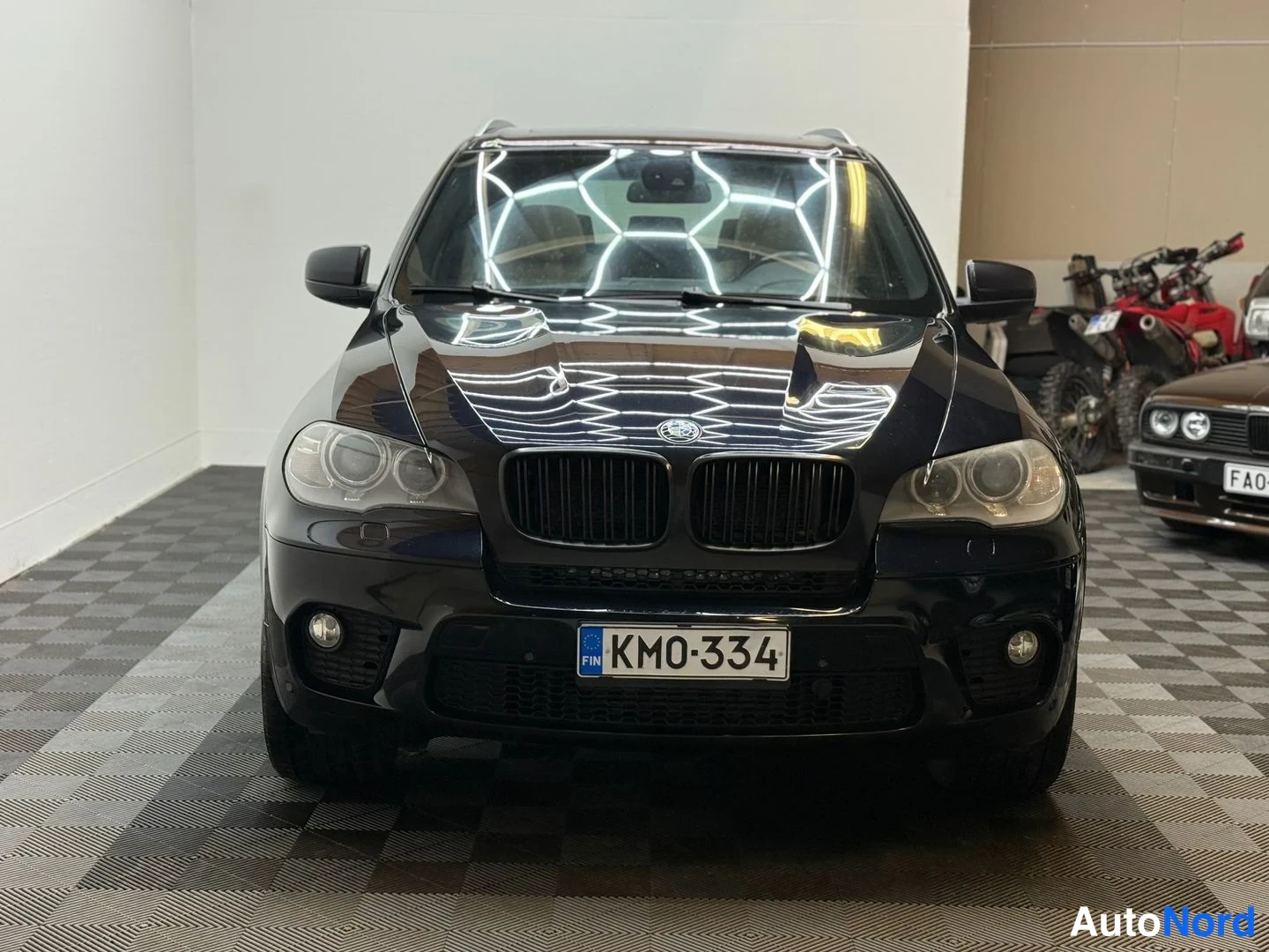 Edestä: Suora etunäkymä mustasta BMW X5:stä sisätiloissa, rekisterikilpi, taustalla toinen auto ja mönkijöitä.